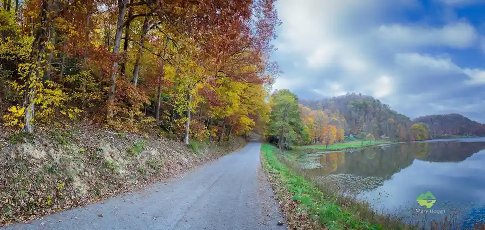 A scenic autumn road winds past a calm lake, flanked by trees with vibrant yellow, orange, and red leaves under a partly…