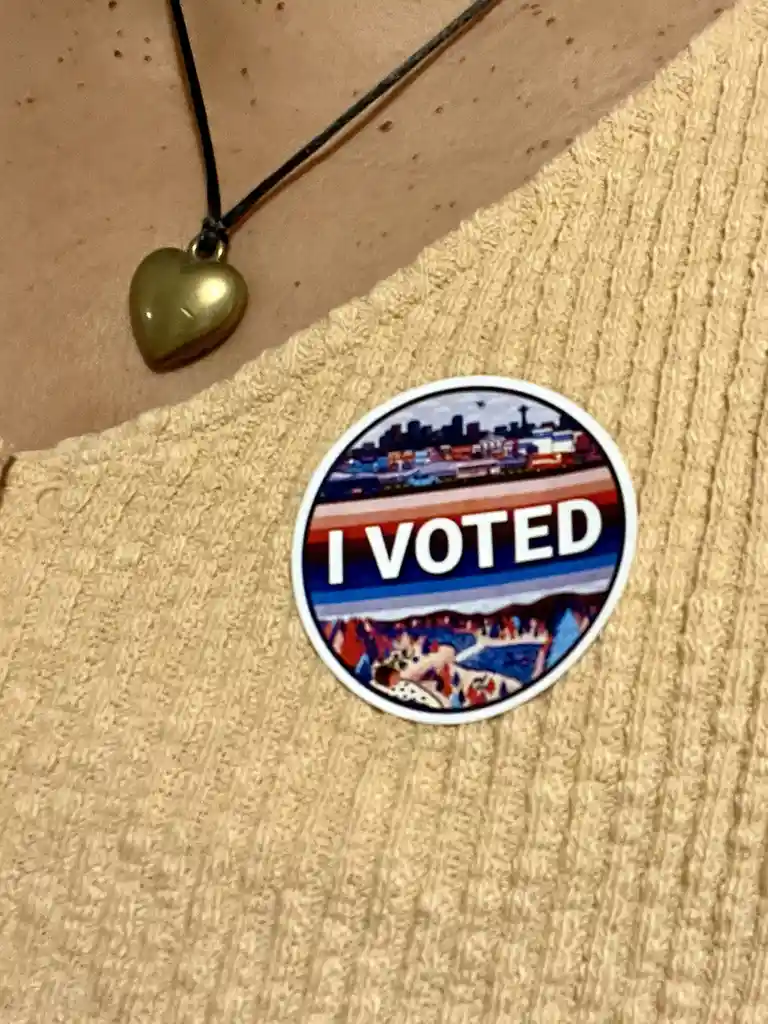 A close-up photo shows a circular 'I VOTED' sticker pinned to a textured beige knit top. The sticker features a stylized…