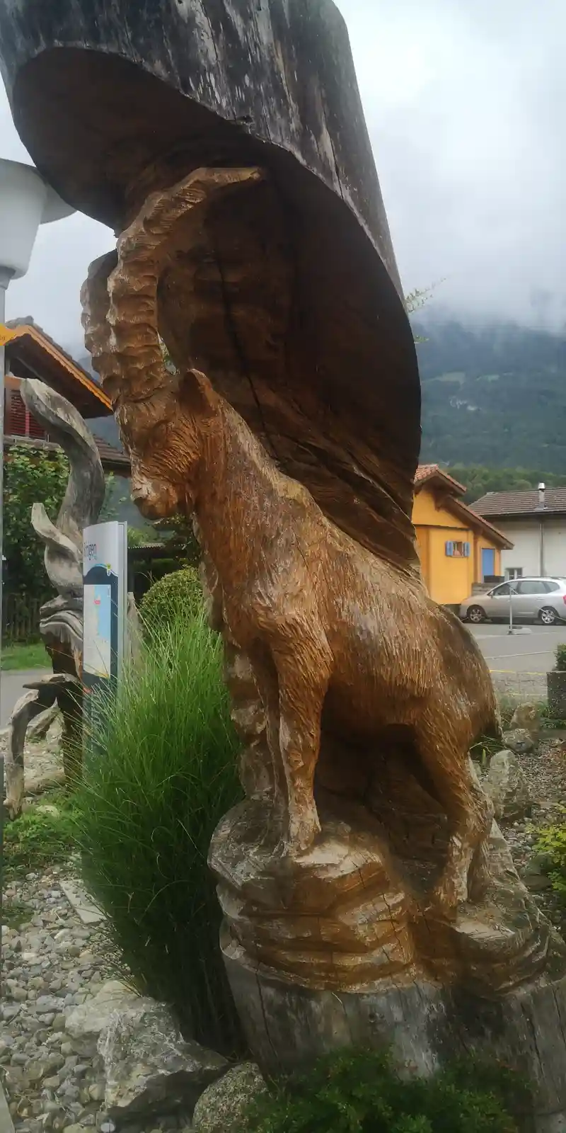 A large, intricately carved wooden sculpture of a goat stands outdoors, its body and head detailed with visible wood grain…