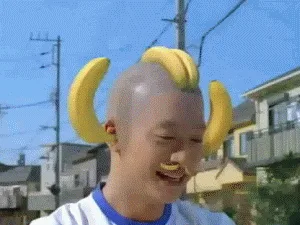 A man with bananas growing out of his head and mustache, laughing and holding a banana, while a woman sits nearby holding a…