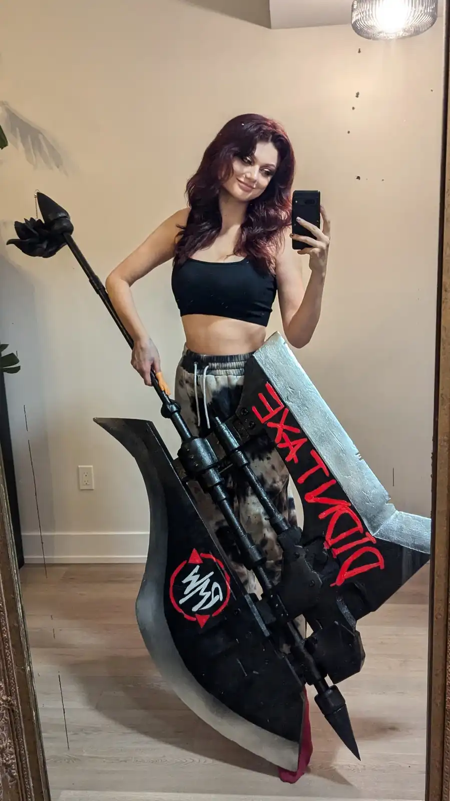 A woman with long dark hair takes a mirror selfie while holding a large, custom-made prop weapon resembling a giant axe or…
