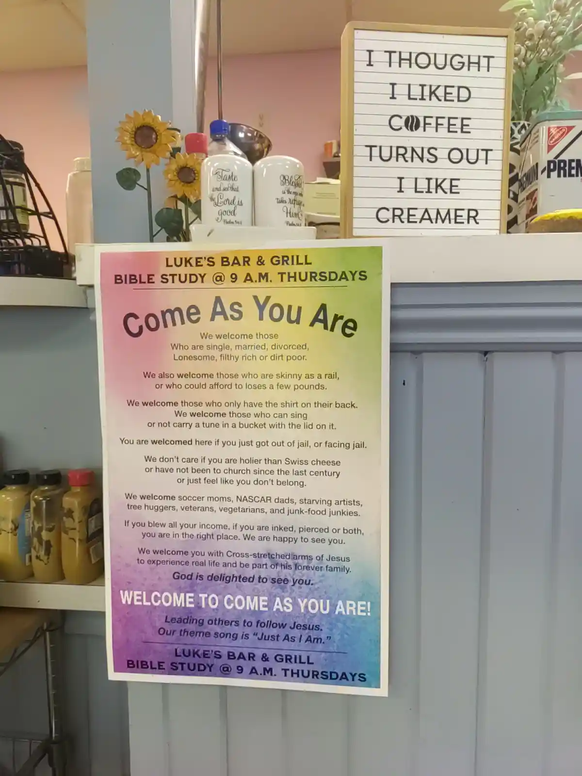 A sign in a cafe reads 'I THOUGHT I LIKED COFFEE TURNS OUT I LIKE CREAMER' above a colorful poster for 'LUKE'S BAR & GRILL…
