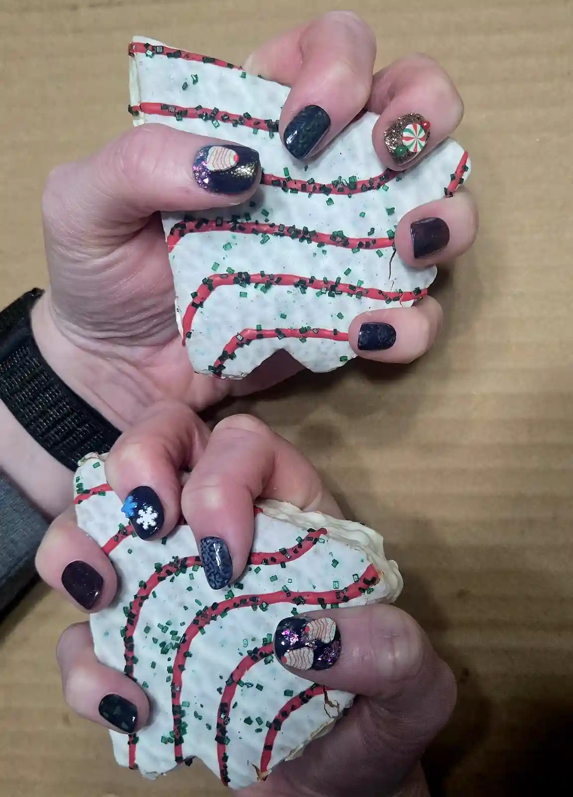 Two hands with festive holiday-themed nail art hold two large, white-frosted cookies decorated with red icing lines and…