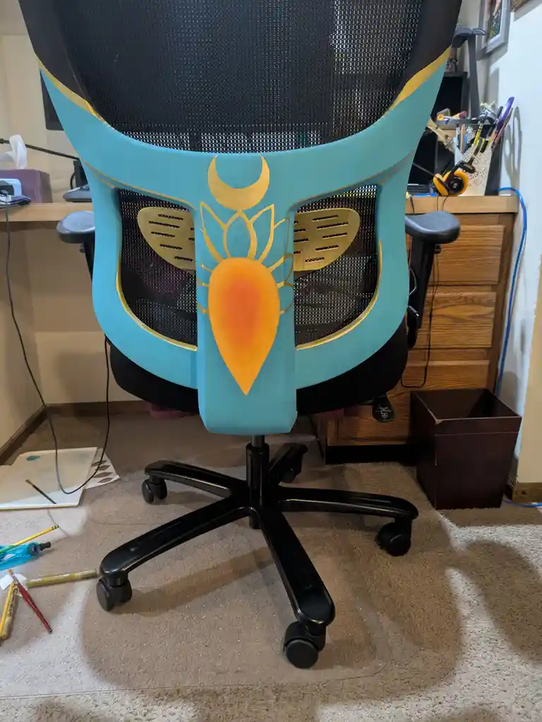 A black office chair has been painted turquoise and decorated with a golden mask-like design featuring a crescent moon…