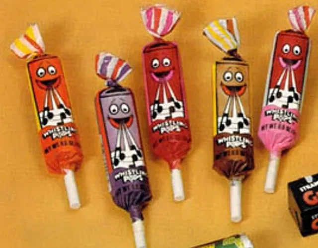 Five Whistlin' Dixie lollipops with cartoon faces and checkered patterns are arranged on a yellow surface. Each lollipop has…