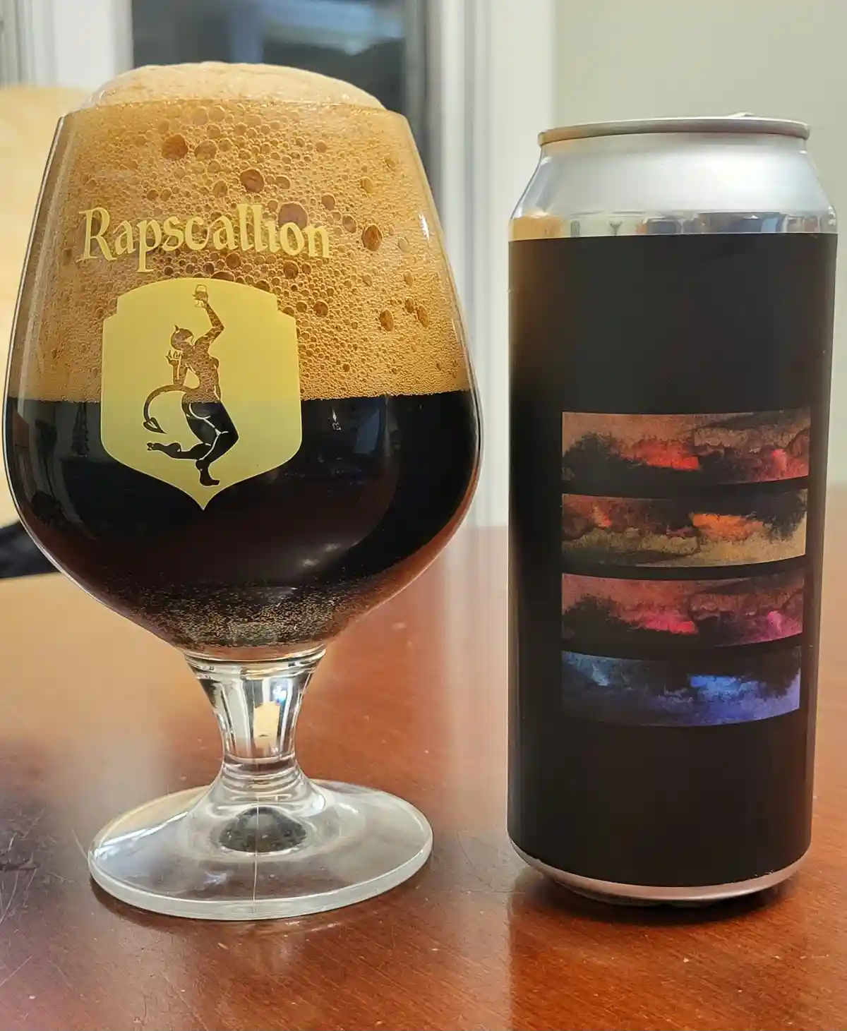 A dark beer in a stemmed glass with a thick, foamy head sits next to its can, which has a black label with four horizontal…
