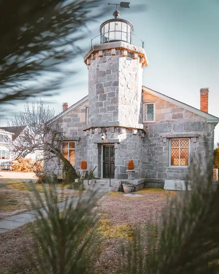 A picturesque stone lighthouse building with a lantern room and weather vane, surrounded by grass and trees, with overlaid…