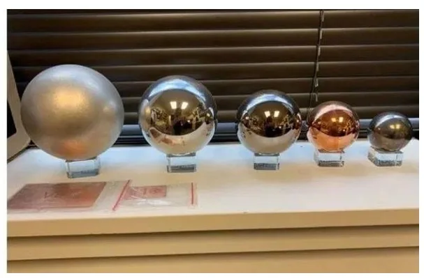 Five polished metallic spheres of decreasing size are arranged in a row on clear stands atop a white surface, with a window…