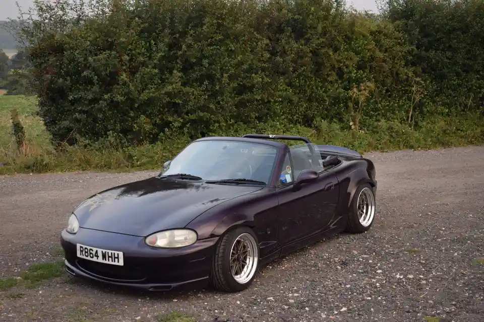A dark purple Mazda MX-5 Miata convertible with aftermarket wheels and a roof rack is parked on a gravel road beside a dense…