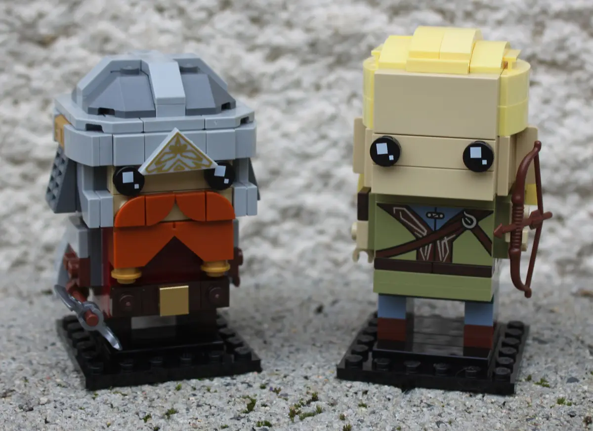 Two blocky, LEGO-style figurines stand side by side on gravel. The figure on the left wears a gray helmet with a golden…