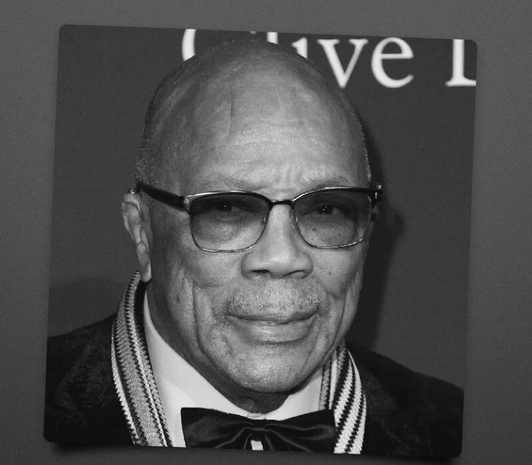 A black and white photo of an older man with glasses, wearing a bow tie and a patterned scarf, looking directly at the…