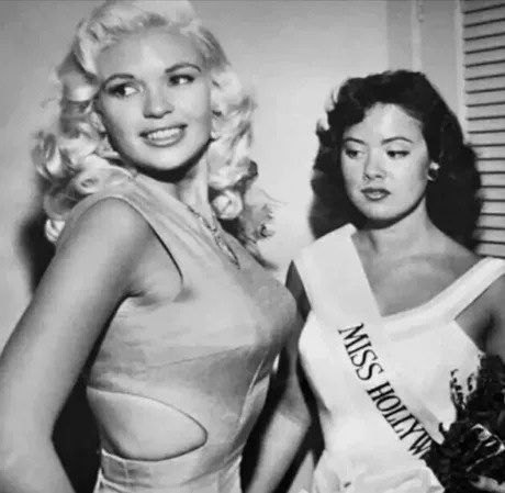 A black-and-white photo shows two women posing together. The woman on the left, with blonde curls and a glamorous dress…