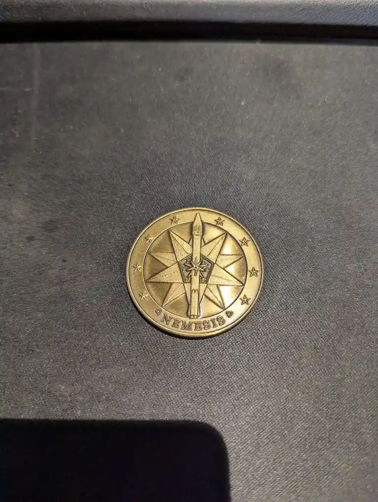 A golden circular token with a rocket ship centered on a starburst design, surrounded by eight stars and the word 'NEMESIS'…