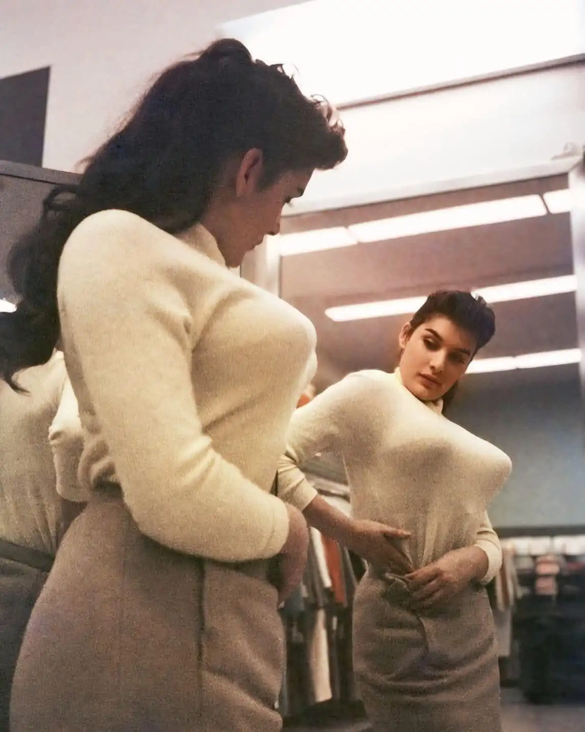 A woman in a cream-colored sweater and skirt stands in front of a mirror, adjusting her clothing while looking at her…