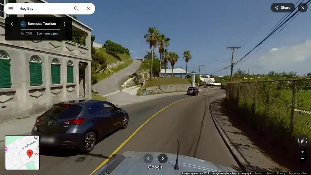 A google street view image from Bermuda showing a car driving down a winding road past a building with green shutters, with…