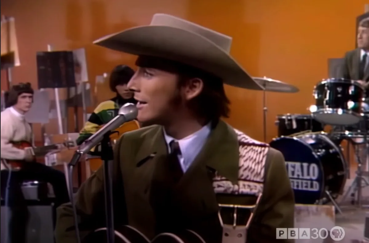A man in a cowboy hat and green jacket sings into a microphone while holding a guitar, with a band behind him. A drum set…
