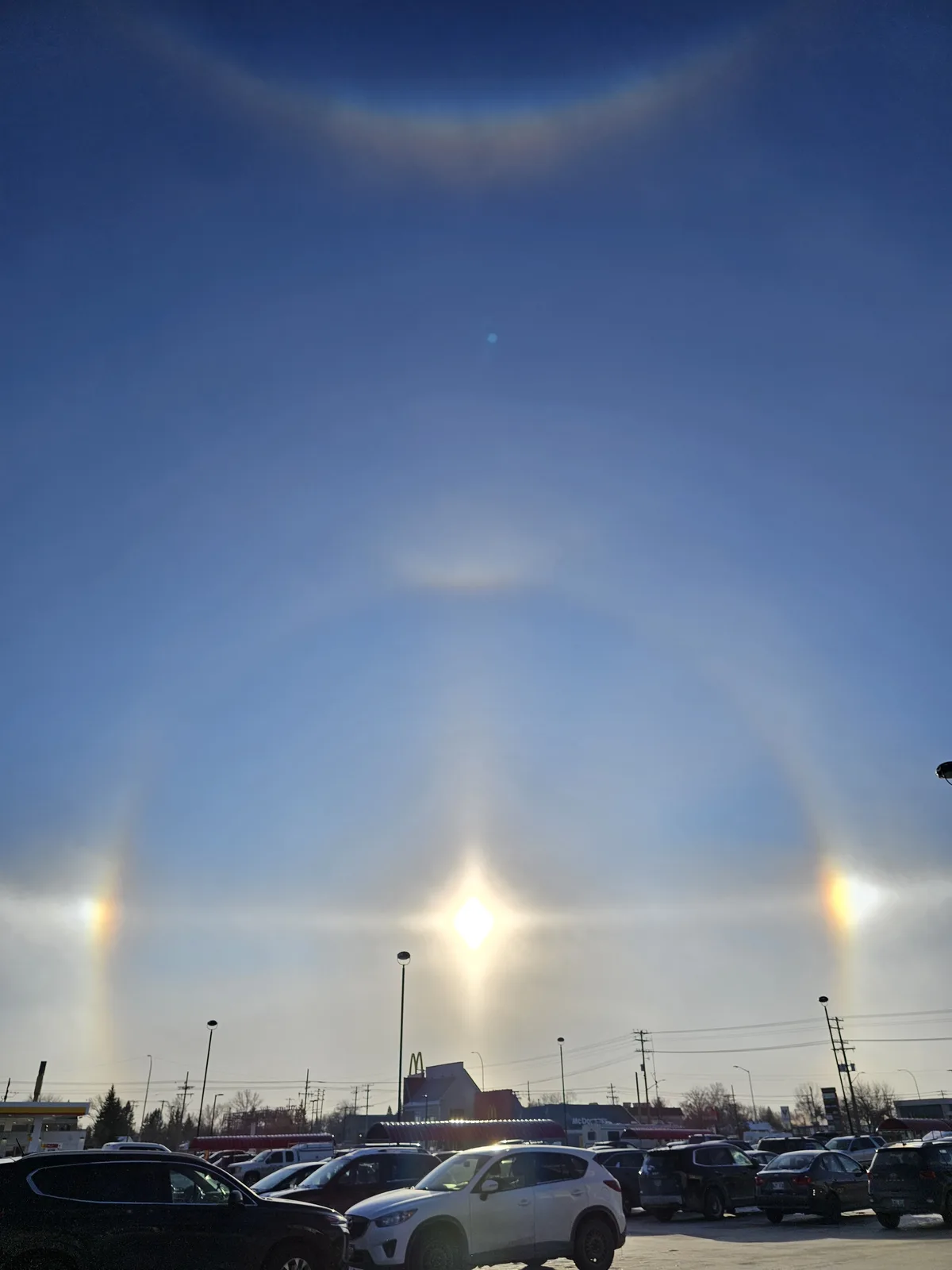 A photograph of a bright sun in a clear blue sky, surrounded by a large, faint circular halo and two smaller halos on either…