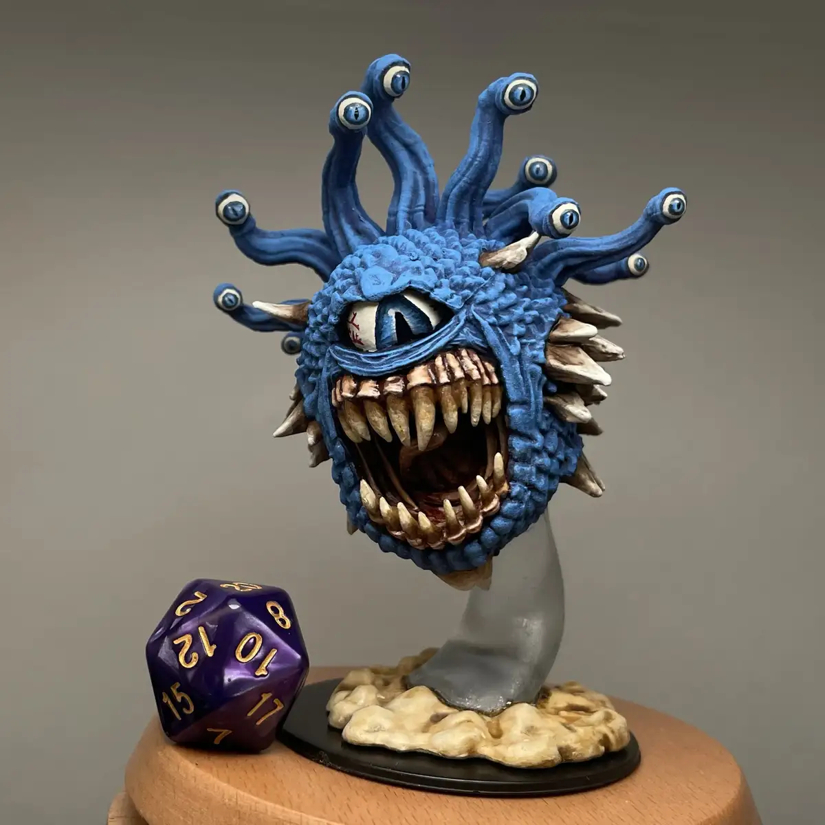 A detailed blue monster figurine with multiple eye stalks and sharp teeth sits on a stand next to a purple 20-sided die…