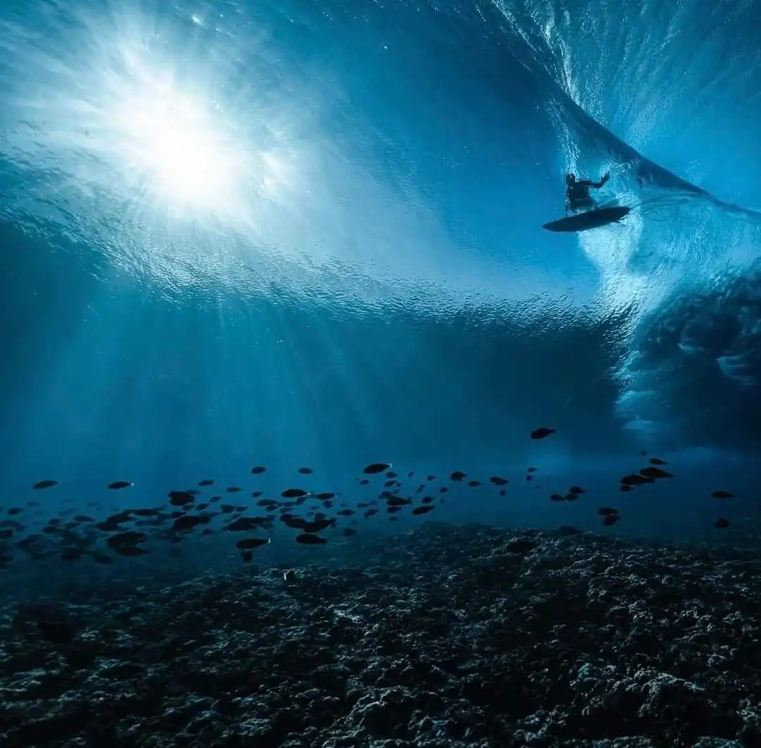 An underwater photograph captures a surfer riding inside a massive, curling ocean wave, with sunlight streaming down from…