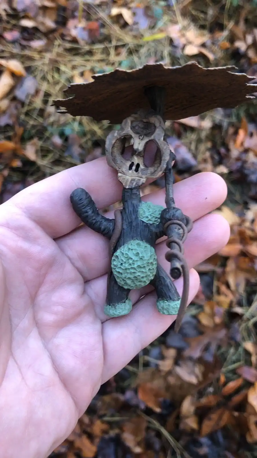 A hand holds a small, eerie figurine crafted from natural materials like bark and acorns, resembling a skeletal creature…