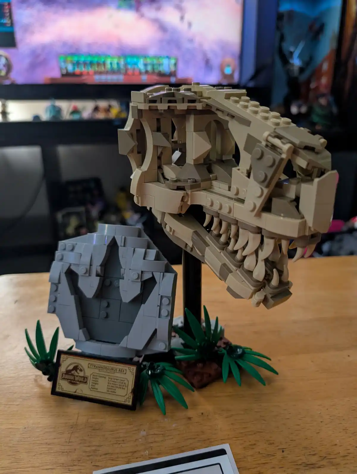 A detailed LEGO model of a Tyrannosaurus Rex skull, mounted on a stand and displayed on a wooden table with small green LEGO…