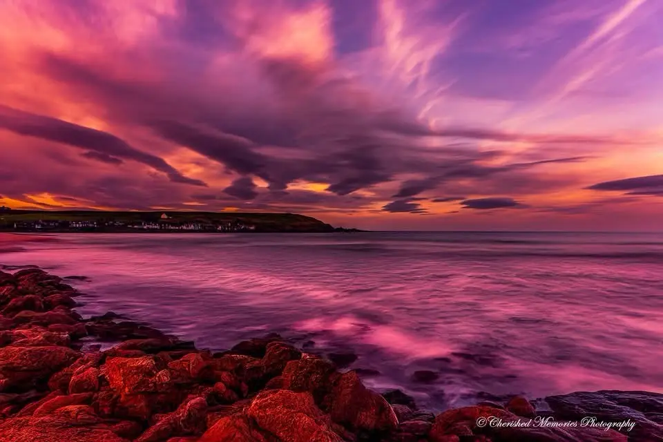 A long-exposure photograph of a vibrant pink and purple sunset over a rocky coastline with calm water, overlaid with the…
