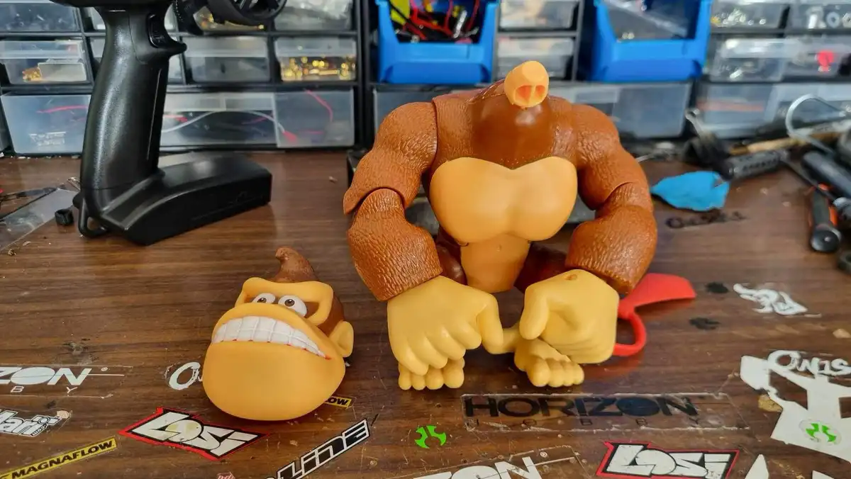 A large, muscular Donkey Kong action figure sits on a cluttered workbench next to a small, separate Donkey Kong head with a…