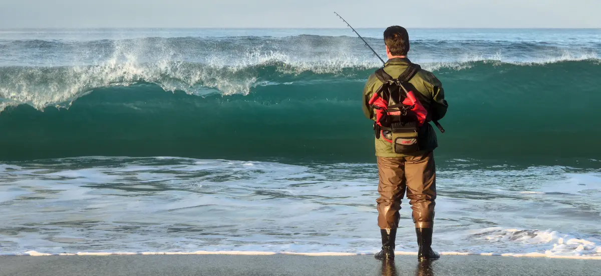 A man in waders and a backpack stands on a beach facing a large, crashing ocean wave while holding a fishing rod. The…