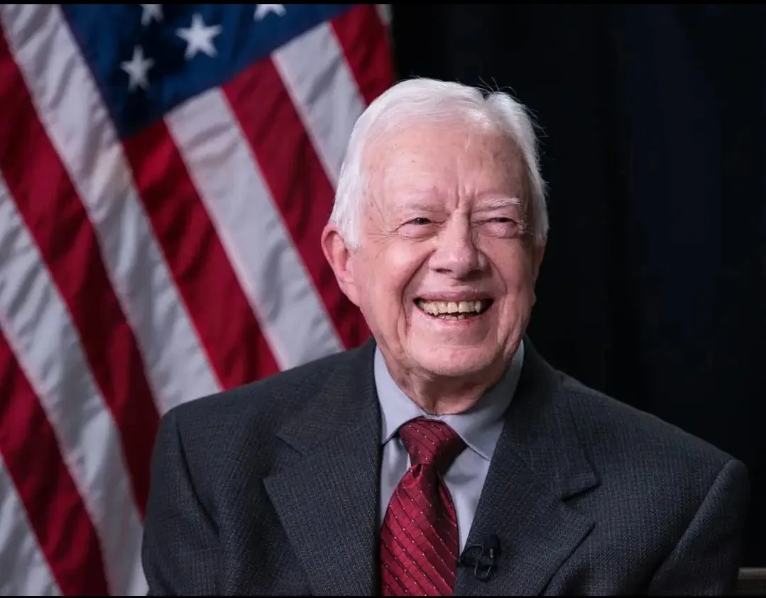 An older white man with white hair, wearing a dark suit and red tie, smiles broadly while seated in front of an American…