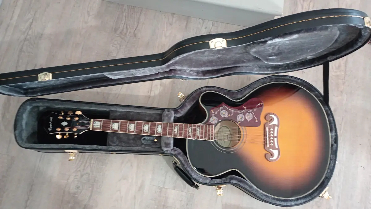 An Epiphone acoustic guitar with a sunburst finish and ornate inlays rests inside its open black hardshell case on a light…