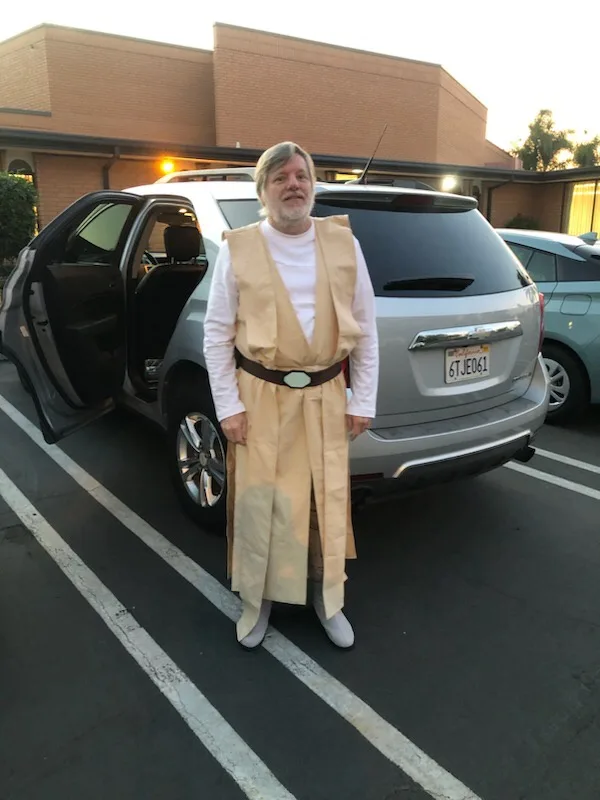 A man dressed in a beige Jedi robe and belt stands in a parking lot next to a silver SUV with its driver's door open. The…