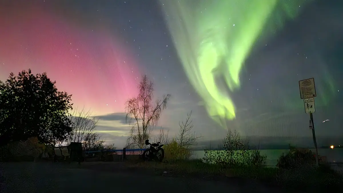 A nighttime photo shows vibrant green and pink auroras dancing across a starry sky above a roadside scene with trees, a…