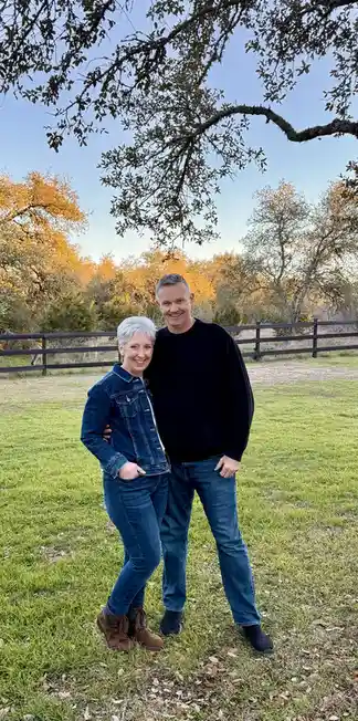 A smiling older couple poses together in a grassy field under a tree with autumn-colored trees in the background. The woman…