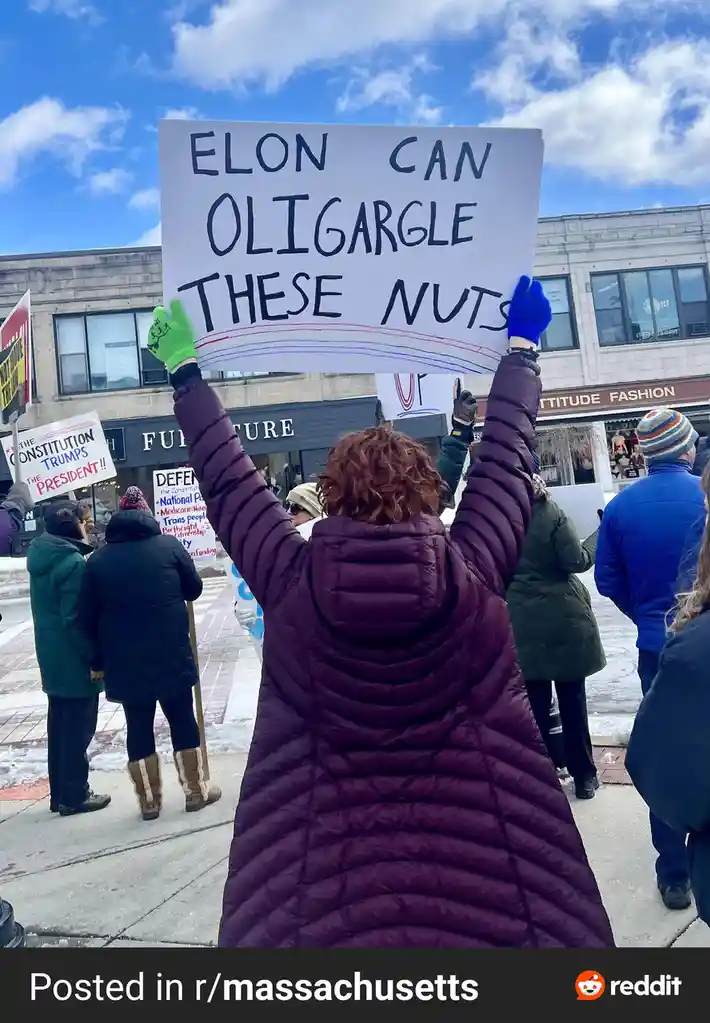 A person in a purple puffer jacket and colorful gloves holds up a handmade protest sign that reads 'ELON CAN OLIGARGLE THESE…