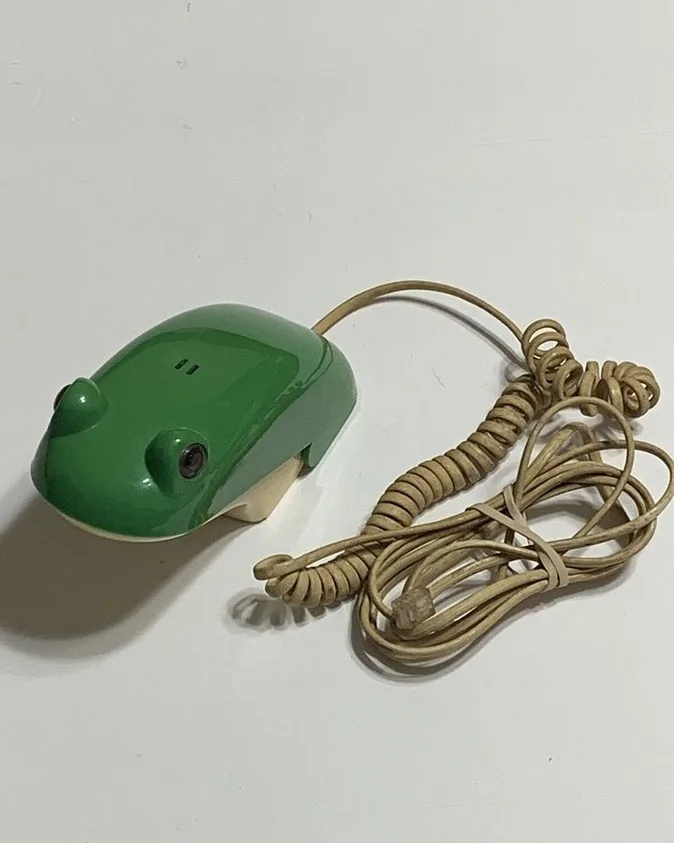 A green, frog-shaped computer mouse with a coiled beige cable lies on a plain white surface. The mouse has molded frog…