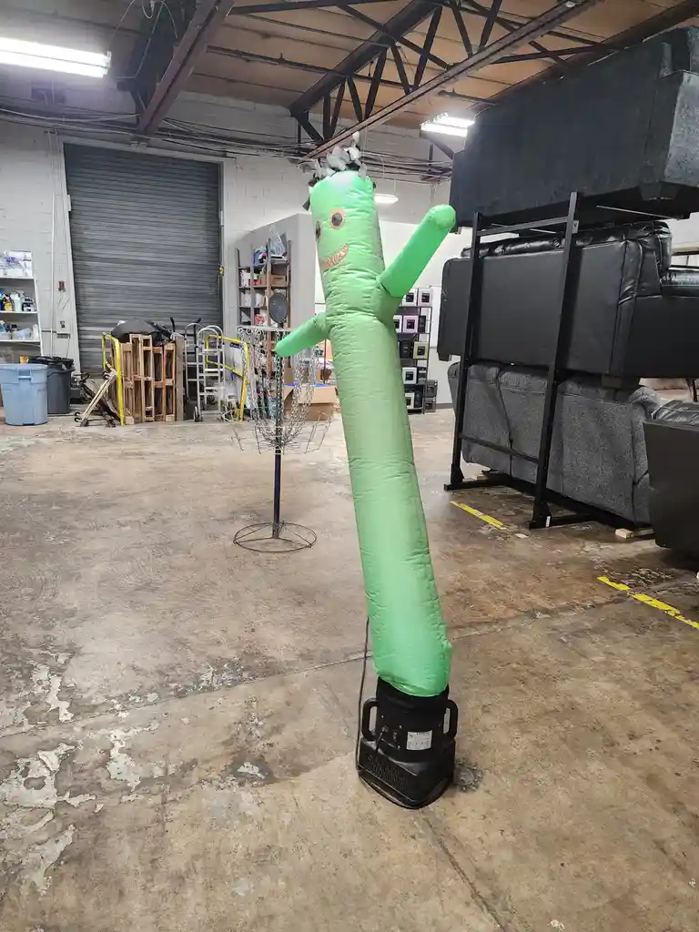 A green inflatable tube man with a smiling face and arms raised stands in a warehouse, its head tangled in wires and debris…