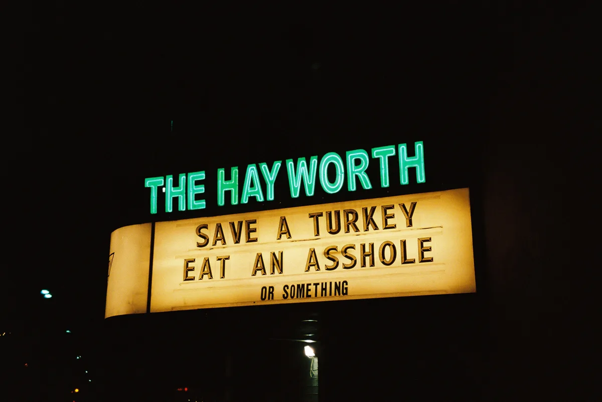 A nighttime photo of a theater marquee for 'THE HAYWORTH' with a lit-up sign below that reads 'SAVE A TURKEY EAT AN ASSHOLE…