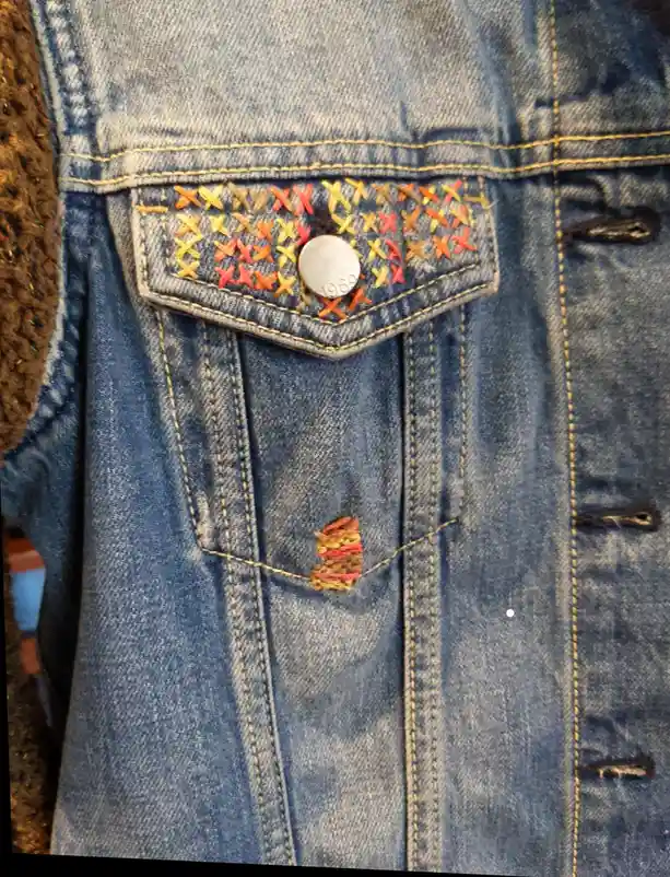 A close-up of a blue denim jacket's chest pocket, which is decorated with colorful cross-stitch embroidery in orange…