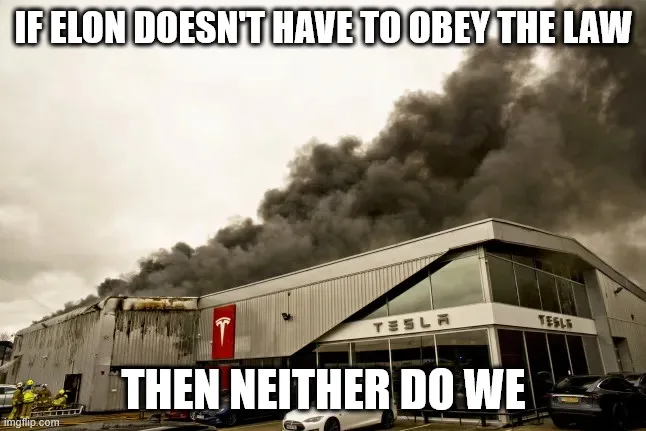 A sepia-toned photo shows a large industrial building with 'TESLA' signage and a red Tesla logo, billowing thick black smoke…