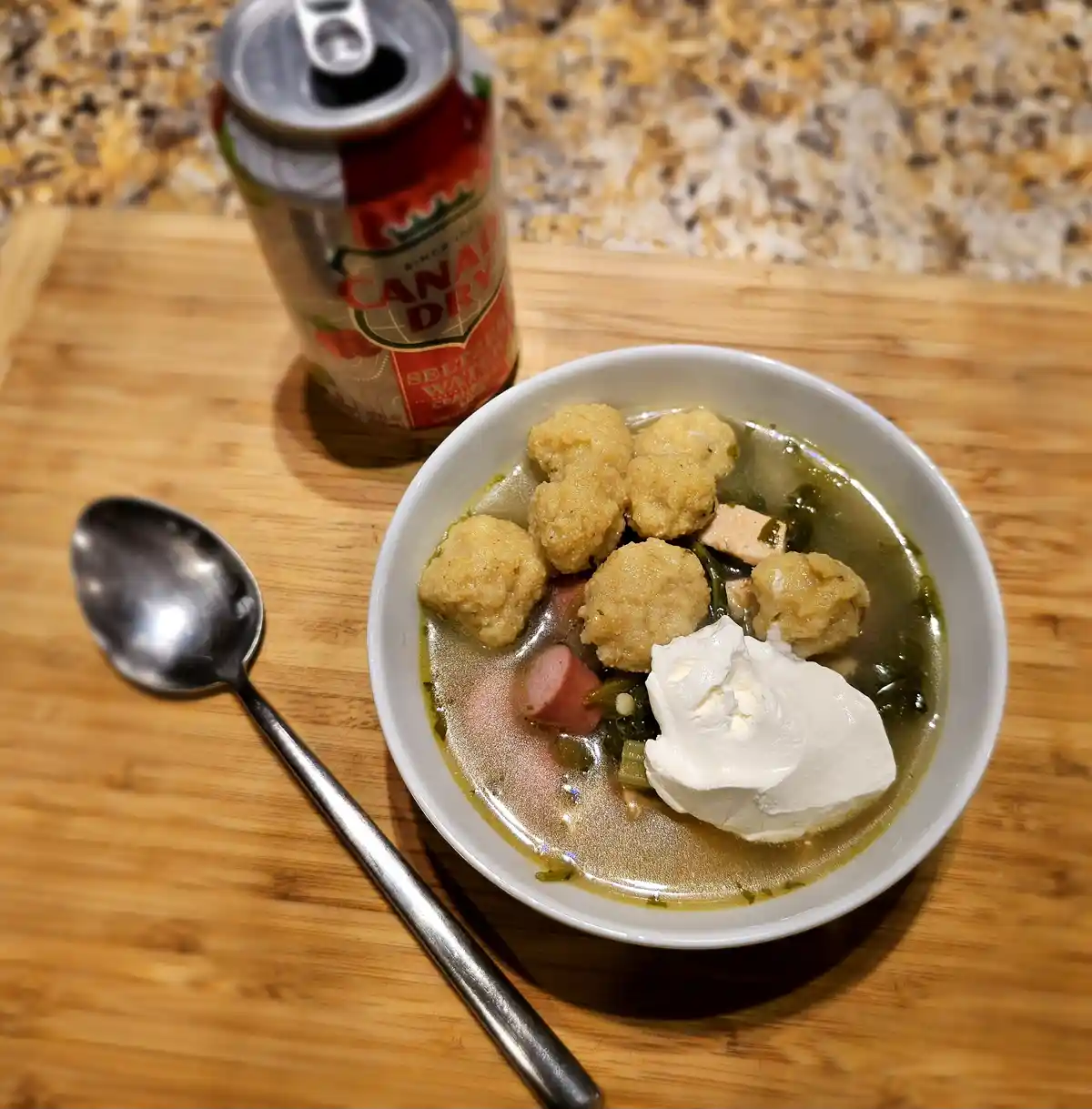 A bowl of soup with dumplings and a dollop of sour cream sits on a wooden cutting board next to a spoon and an open can of…