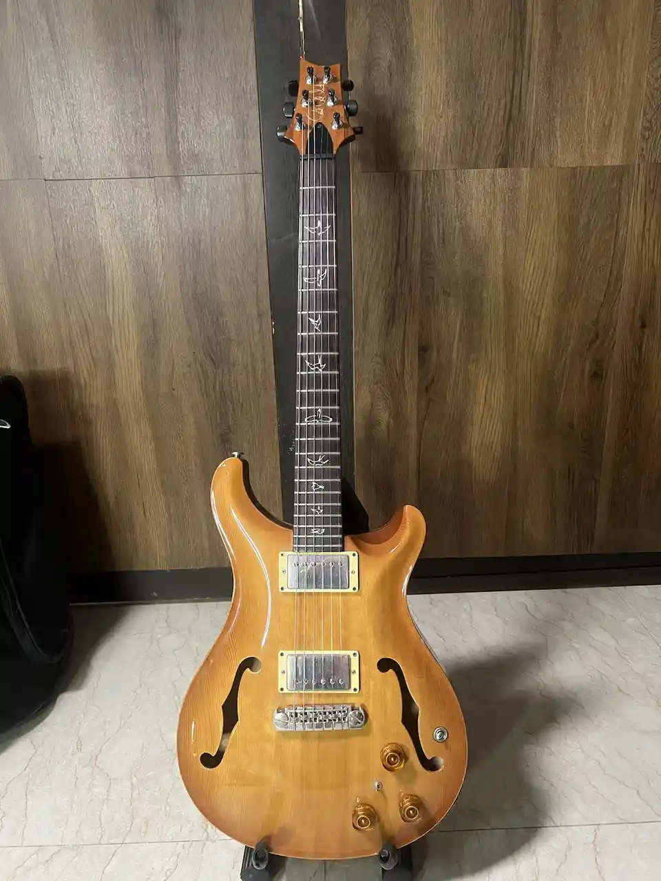 A light-wood-finish electric guitar with f-holes and gold hardware stands upright on a stand against a wood-paneled wall and…
