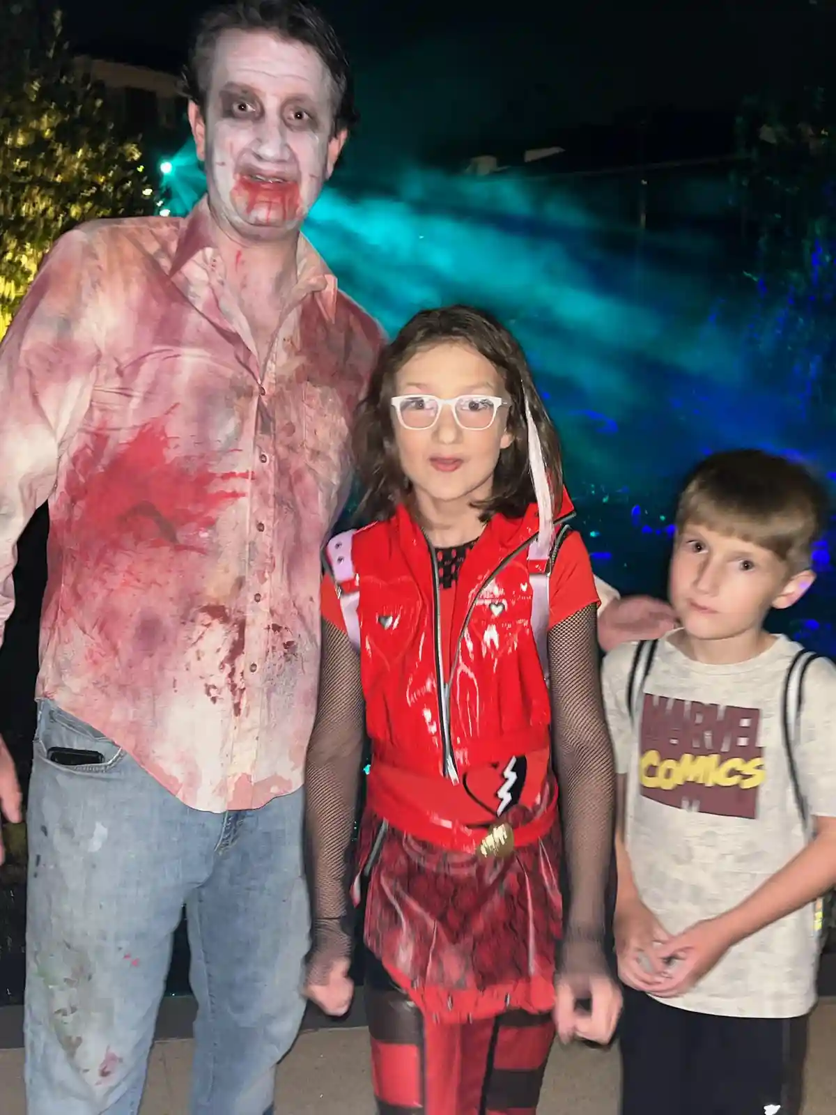 A man dressed as a zombie with fake blood on his face and shirt stands beside two children at night. The girl wears a red…