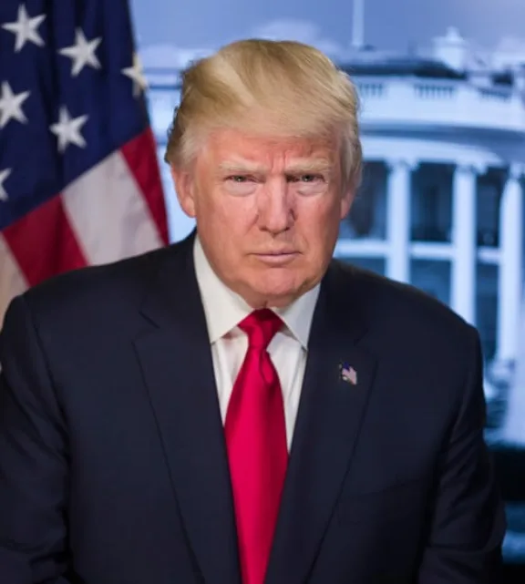 a formal portrait of Donald Trump wearing a dark suit and red tie, standing in front of an American flag and a blurred image…