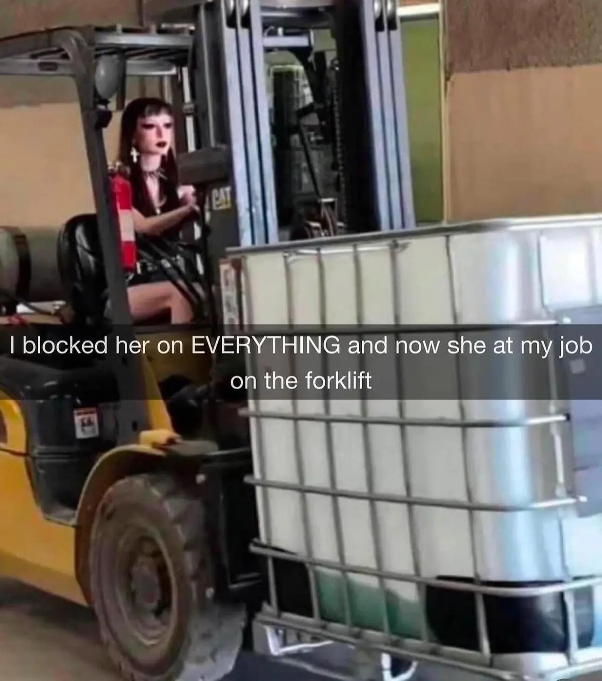A person with dark hair and heavy makeup sits in the driver's seat of a yellow forklift, which is carrying a large metal IBC…