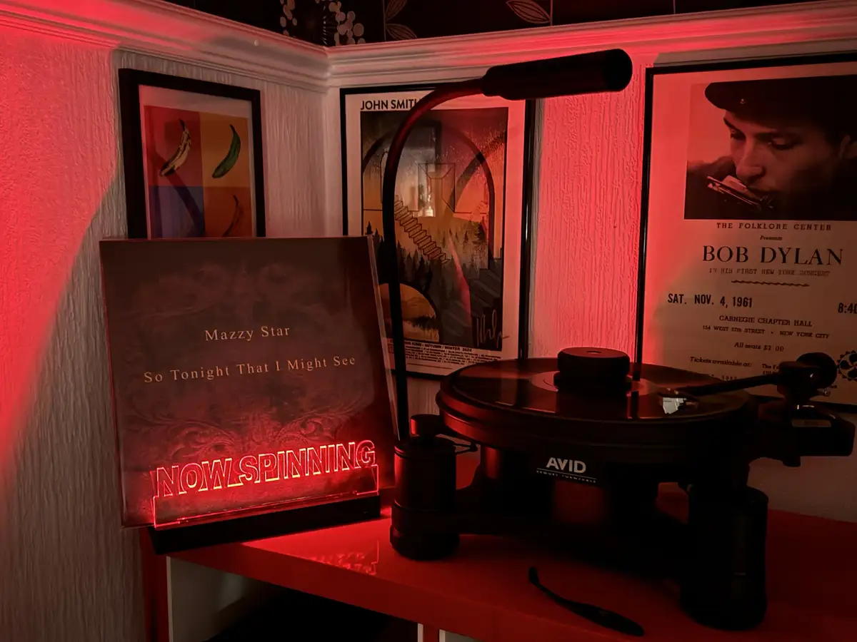 A vinyl record player sits on a red shelf under red lighting, with a glowing sign next to it that reads 'Mazzy Star So…