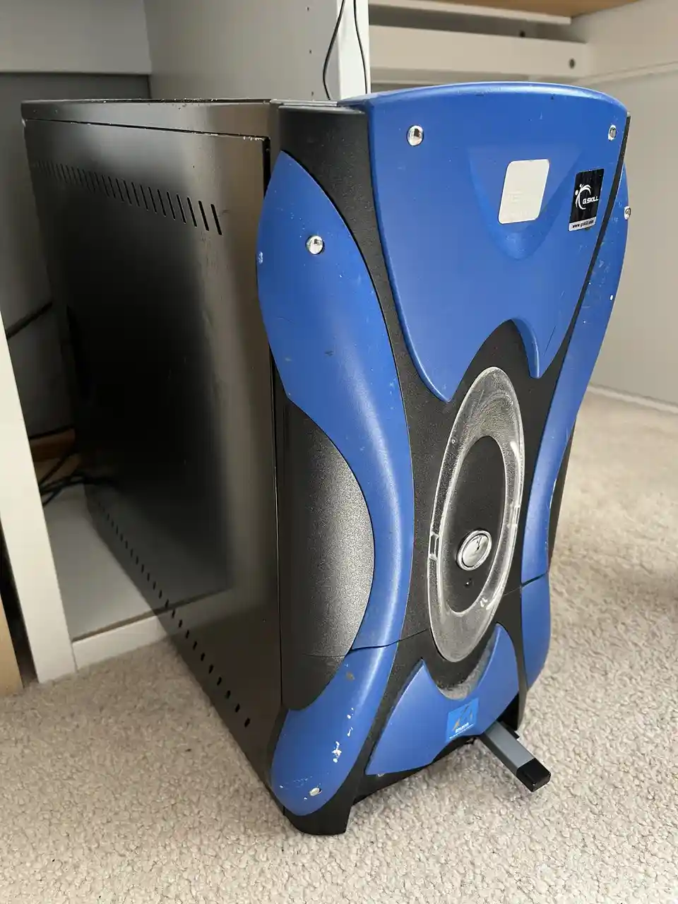 A blue and black computer case with a large circular speaker-like feature on its side sits on a carpeted floor, visibly…