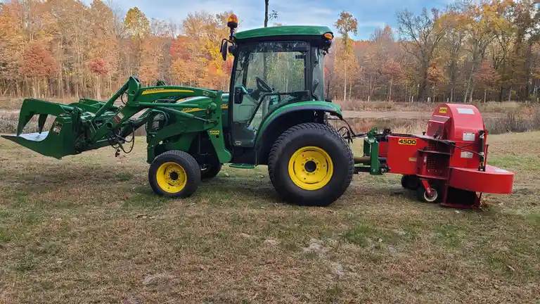 A green John Deere 4720 tractor with a front loader is parked on grass near a pond, with autumn trees in the background…