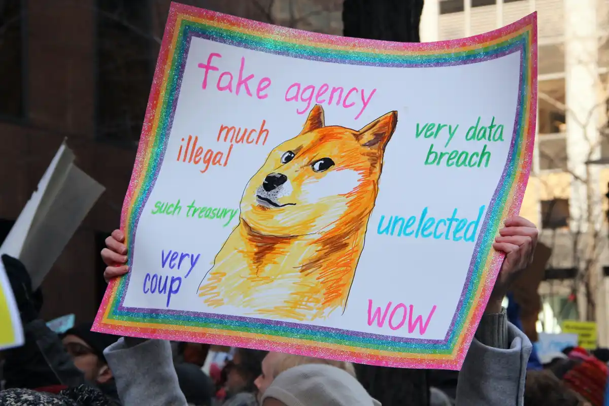 A person holds a handmade protest sign with a rainbow glitter border featuring a drawing of a Shiba Inu dog. Around the dog…