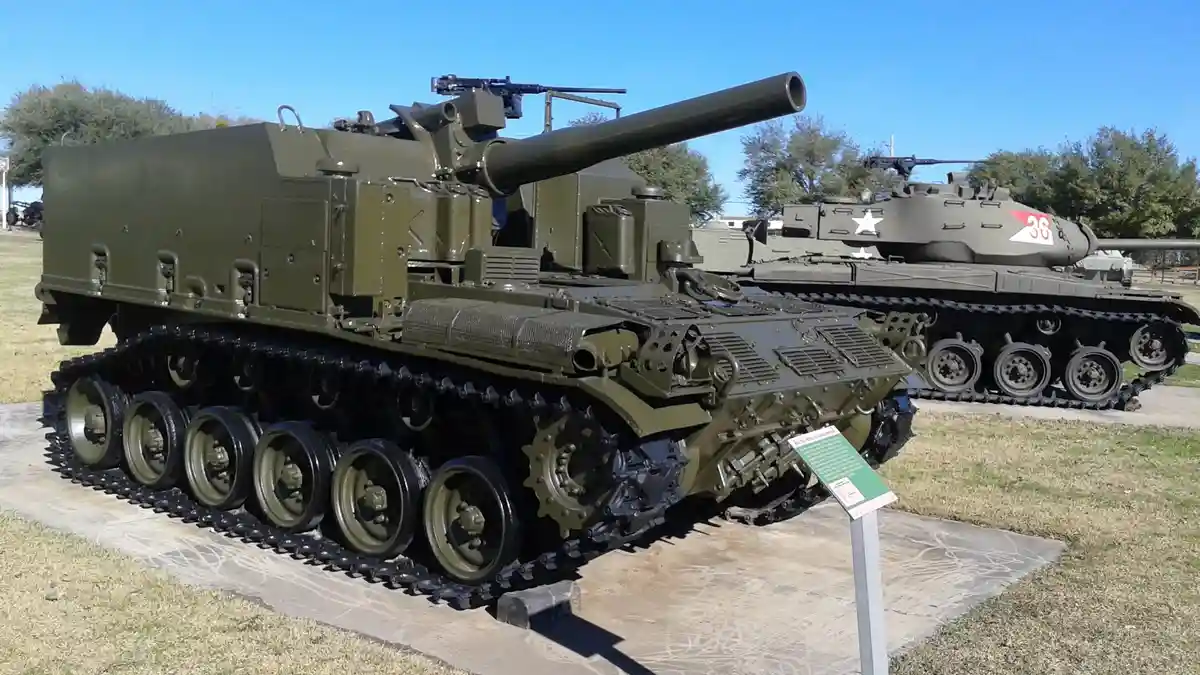 A large olive-drab self-propelled artillery vehicle with a long barrel and tracks is displayed outdoors, with another tank…