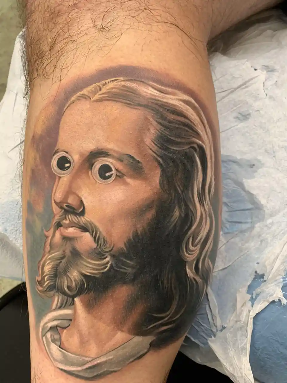 A realistic tattoo of Jesus Christ on a person's leg, featuring detailed shading and a halo, but with cartoonish googly eyes…